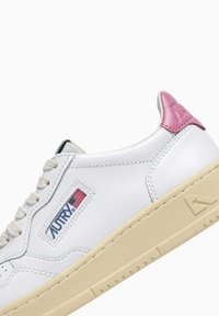 Sneaker in pelle bianca con suola beige testurizzata, dettaglio rosa sul tallone, chiusura con lacci e una toppa con logo blu con una piccola bandiera americana.