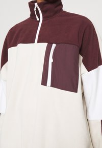 Pull polaire à demi-fermeture éclair avec partie supérieure bordeaux, partie inférieure crème et poche poitrine bordeaux avec fermeture éclair blanche verticale.