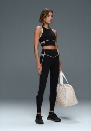 Vrouw draagt een zwart atletisch crop top en legging met witte rand, houdt een beige gestikte sporttas vast, staand tegen een grijze achtergrond.