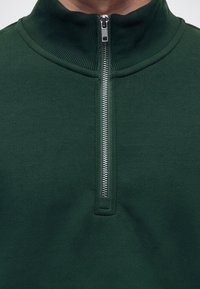 Πράσινο φλις πουλόβερ με ριμπ ύφασμα στη ψηλή λαιμόκοψη και ασημένιο φερμουάρ, μερικώς ανοιχτό στο κέντρο μπροστά.