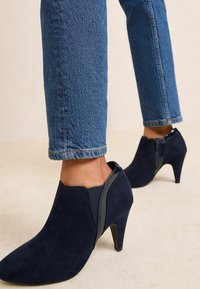 Botas de ante azul marino hasta el tobillo con punta afilada y tacón de altura media. Panel elástico lateral y cremallera para facilitar su uso. Jeans con los puños doblados visibles.