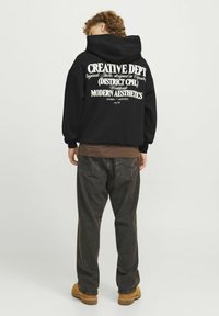 Zwarte hoodie met een groot wit grafisch tekst op de achterkant, met een ontspannen pasvorm en geribbelde manchetten. Gecombineerd met donkergrijze broek en bruine schoenen.