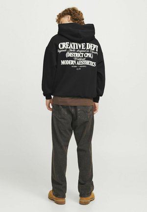 Black hoodie with a large white graphic text on the back, featuring a relaxed fit and ribbed cuffs. Paired with dark gray pants and brown shoes.