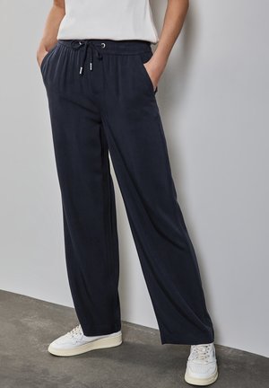 Pantalons bleu marine, ample, avec une taille élastique et un cordon de serrage. Comprend des poches latérales et une texture douce, associés à des baskets blanches.