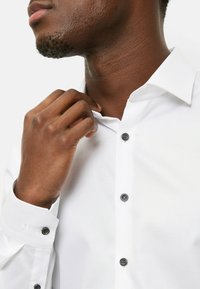 Weißes Hemd mit spitzem Kragen, dunkelblauen Knöpfen und strukturierter Stoffstruktur. Eine Hand richtet den Kragen am Hals.