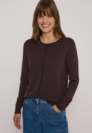 Femme souriante portant un pull à manches longues marron foncé et un jean bleu, une main dans la poche, sur un fond clair uni.