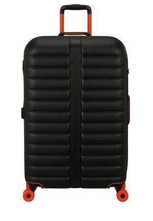 Maleta rígida negra con ranuras horizontales, asa telescópica naranja y cuatro ruedas giratorias naranjas sobre fondo blanco.