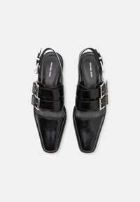 Musta kiiltävästä nahasta valmistetut slingback-kengät, joissa on neliskulmaiset kärjet, kaksi hopeista solkea ja sileä pinta. Tasapohjaiset ja minimalistisella muotoilulla.