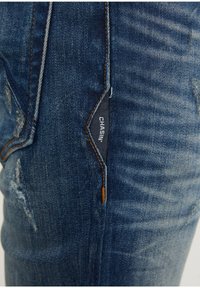 Tummansiniset farkut, joissa kulutuksen jälkiä. Ominaisuuksina "CHASIN'"-logolla varustettu merkki ja kontrastiväriset saumat. Lähikuva sivunäkymästä.