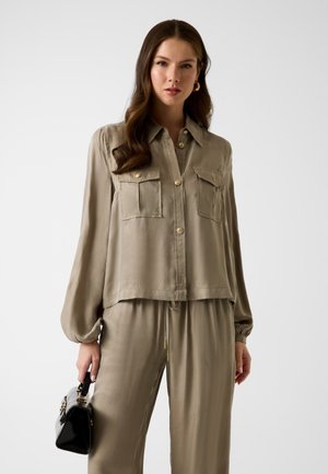 Femme portant une chemise beige à boutons avec poches poitrine et un pantalon assorti, tenant un petit sac à main noir dans sa main gauche.