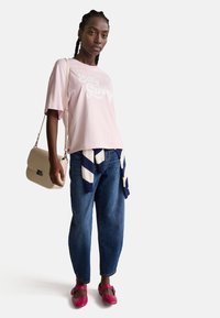 Růžové bavlněné tričko s bílým nápisem "Stay Groovy", spárované s modrými džínami, růžovými botami a béžovou crossbody kabelkou. Pruhovaný námořnický svetr svázaný v pase.