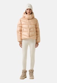 Kurtka puchowa w delikatnym brzoskwiniowym kolorze z wysokim kołnierzem, napami i pikowanym wzorem. Połączona z kremowymi legginsami i beżowymi skórzanymi butami podszewką z futra.