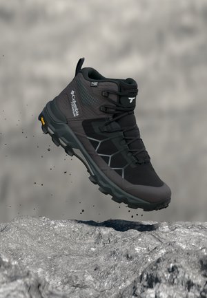 Schwarzer und grauer Columbia Titanium Wanderschuh mit Vibram-Sohle in der Luft über felsigem Gelände, zeigt Seiten- und Vorderansicht.