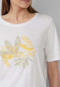 T-shirt à manches courtes blanc avec un graphisme "STAY VACAY" en jaune et gris agrémenté de motifs de feuilles. Tissu doux et texturé avec une coupe décontractée.