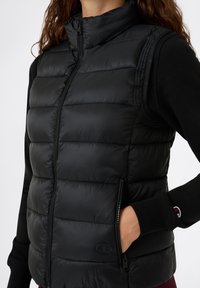 Gilet imbottito nero con colletto alto, frontale con zip, tasche laterali e dettagli con logo. Realizzato in materiale liscio e lucido con una texture trapuntata.