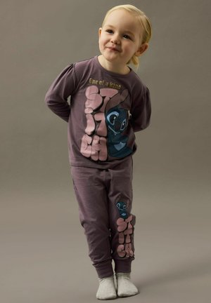 Bambino piccolo in piedi con le mani dietro la schiena, indossa un pigiama viola con il personaggio Stitch e la scritta "Unico nel suo genere".