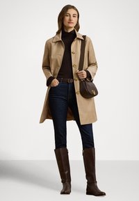 Beige trenchcoat med brun vertikal rand, guldfärgade knappar; mörk polotröja, mörka jeans, bruna läderstövlar upp till knäna och en axelremsväska.