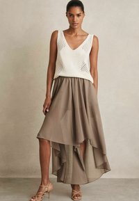 Top in maglia crema con scollatura a V e lati a tessitura aperta, abbinato a una gonna asimmetrica marrone con volants stratificati e un design fluido.