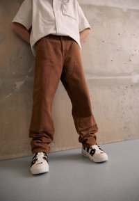 Bruna baggy byxor med synliga sömmar och fickor, kombinerat med vita och marinblå sneakers med en framträdande logotyp på sidan.