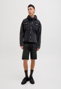 Zwarte denim jack met drukknopen, twee borstzakken, gecombineerd met zwarte denim shorts, donkere sokken en zwarte schoenen. Neutrale achtergrond.