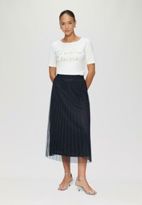 Witte blouse met korte mouwen, voorzien van metallic letters, gecombineerd met een geplooide marineblauwe rok. Zilveren sandalen met bandjes maken de look compleet.