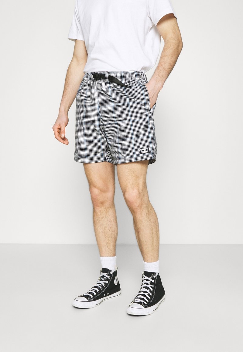 Homme portant un short à carreaux noir et blanc avec une ceinture à boucle noire, des chaussettes blanches montantes, des baskets montantes noires et un t-shirt blanc.