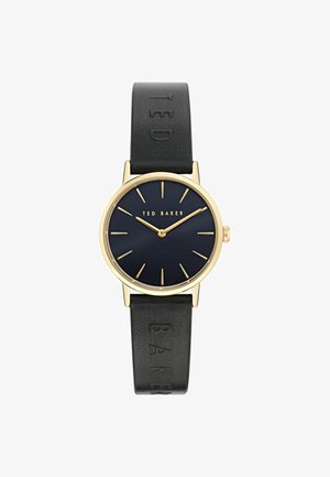 Ümara kujuga kell, millel on kuldne korpus, tumesinine kellanägu, kullast tunni märgid ja must nahkrihm, millel on reljeefne tekst "TED BAKER".