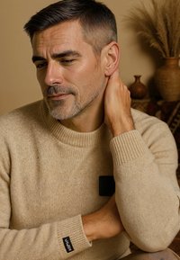 Beige gebreide trui met ribbelboorden en ronde hals. Bevat een zwarte patch op de borst. Soepele textuur met een losse pasvorm.