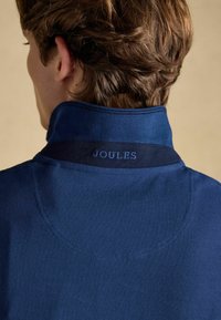 Personne aux cheveux bruns courts portant une veste bleue à col avec "JOULES" brodé sur l'arrière du col.