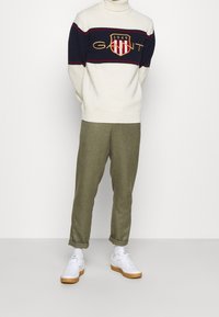 Crèmekleurige gebreide trui met marineblauwe en rode horizontale strepen, voorzien van het "GANT" logo en schild. Olijfgroene broek en witte sneakers met gumzolen.