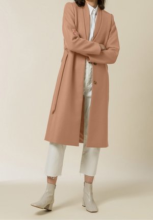 Personne portant un long manteau camel par-dessus un pantalon blanc raccourci et des bottines blanches, debout, les bras croisés devant un fond uni.