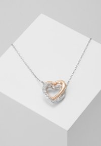 Naszyjnik w kształcie serca z różowym złotem wewnątrz srebrnej warstwy zewnętrznej, ozdobiony przezroczystymi kryształami, zawieszony na delikatnym łańcuszku.