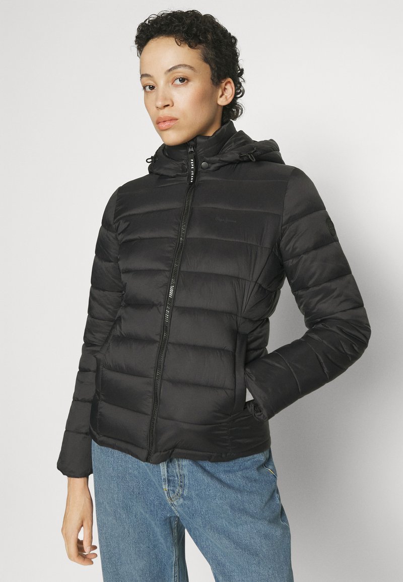 Chaqueta de plumas negra con capucha, frente con cremallera y diseño acolchado. Presenta una silueta ajustada y bolsillos laterales. Lleva puesta sobre jeans de mezclilla.