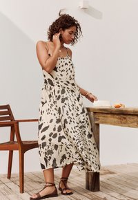 Vrouw in een zwart-wit gestippelde zomerse jurk en bruine sandalen, staand bij een houten tafel met een open boek en een gesneden sinaasappel.