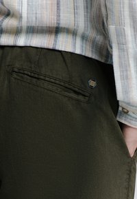 Donkergroene broek van een gestructureerde stof, met zijzakken en een kleine hexagonale metalen accent nabij de taille. Gestreepte shirt zichtbaar erboven.
