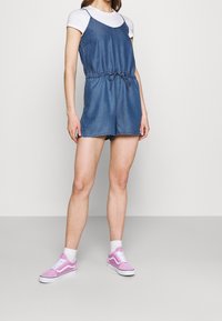 Dżinsowe szorty ogrodniczki z regulowanymi ramiączkami, z elastycznym pasem i luźnym krojem, w zestawie z białą koszulką i różowymi sneakersami.