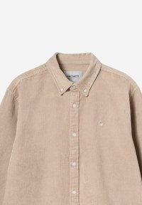 Beżowa koszula z materiału kordowego zapinana na guziki, z kołnierzykiem, białymi guzikami i małym haftowanym logo na piersi poniżej kołnierzyka.