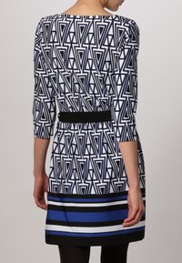 Sukienka z rękawem sięgającym do trzech czwartych w geometryczny czarno-biały wzór, z dopasowaną talią ozdobioną czarnym paskiem i w paskowaną niebieską falbaną u dołu.
