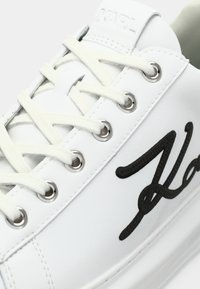Close-up de um ténis branco com atacadores creme, ilhós prateados, "Karl" em relevo na lingueta e um logótipo em script preto na lateral.