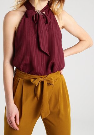 Bordowy top bez rękawów w pionowe paski z wiązaniem przy szyi, zestawiony z musztardowymi spodniami z wysokim stanem i ozdobną kokardą z przodu.