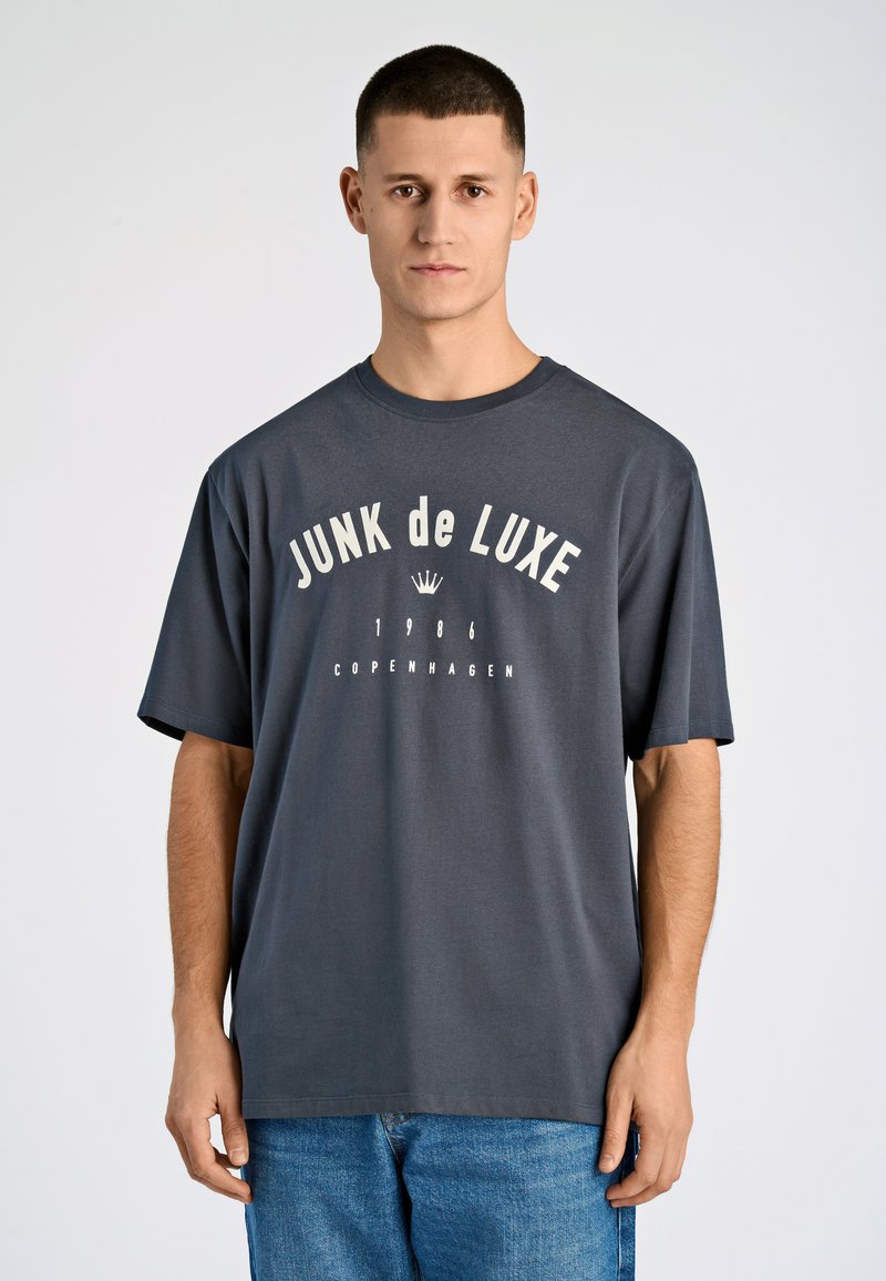 Camiseta de algodón gris con cuello redondo, que presenta texto impreso en blanco: "JUNK de LUXE 1986 COPENHAGUE." Mangas cortas y ajuste relajado.