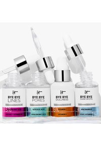 Quatre sérums dermiques concentrés IT Bye Bye étiquetés Lignes, Pores, Teint Terne, Taches Sombres avec des bouchons à pipette sur une surface blanche.