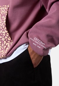 Purple sweatshirt with a leopard print pocket, featuring a ribbed texture and branding on the sleeve. Worn over a white shirt.