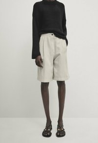 Musta neulepusero leveillä hihoilla, beige räätälöidyt shortsit etutaskuilla ja litteät mustat sandaalit kultaisilla yksityiskohdilla.