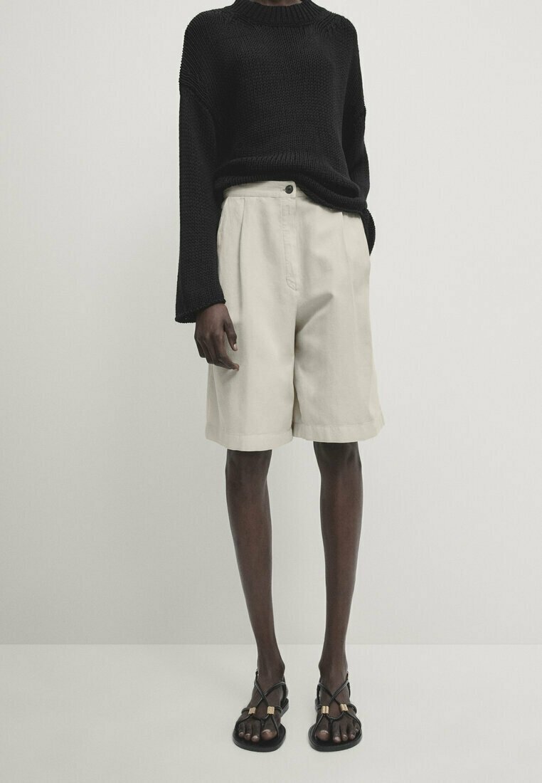 Musta neulepusero leveillä hihoilla, beige räätälöidyt shortsit etutaskuilla ja litteät mustat sandaalit kultaisilla yksityiskohdilla.