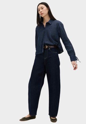 Kvinde med langt mørkt hår iført en løs mørk denimskjorte, højtaljede viddeben mørke jeans, brun bælte og olivengrønne balletsko.