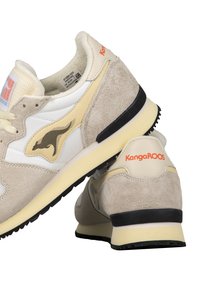 Scarpe da atletica beige e bianche realizzate in materiale scamosciato e mesh, con suole nere e un logo prominente in nero e arancione.
