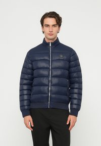 Férfi sötétkék, steppelt pufi dzsekiben, cipzárral és fekete nadrágban, egy egyszerű világos háttér előtt áll.