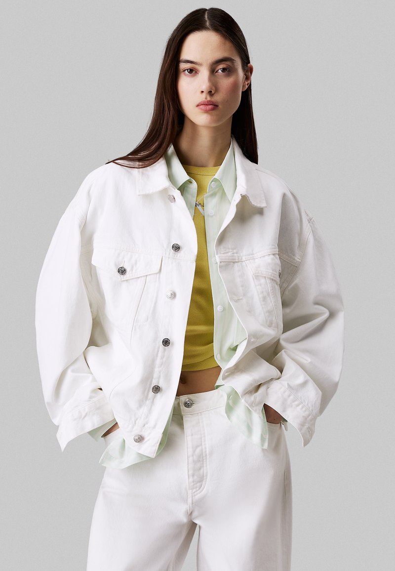 Chaqueta de denim blanca de corte oversize, con botones a presión plateados, superpuesta sobre una camiseta amarilla acanalada y una camisa verde claro con cuello. Pantalones blancos.