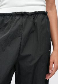 Κοντινό πλάνο ενός ατόμου που φοράει μαύρο ελαφρύ παντελόνι με ελαστική μέση και λευκό τοπ, δείχνοντας μέρος του δεξιού του χεριού.